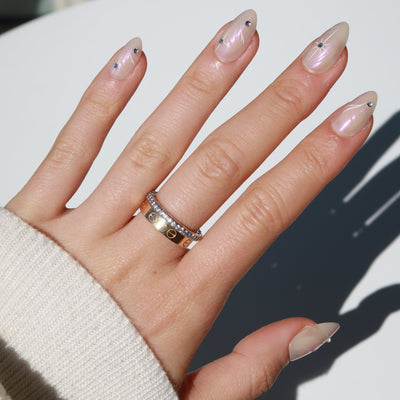 A hand displays salon-quality, almond-shaped nails in the stunning iridescent pink chrome of Glowie By Her's Aura collection, complete with star embellishments. The index and middle fingers are adorned with elegant gold and diamond rings, while a cozy beige sweater sleeve gently wraps around the wrist. Every detail of Aura is completely customizable, allowing for a personalized touch of luxury.