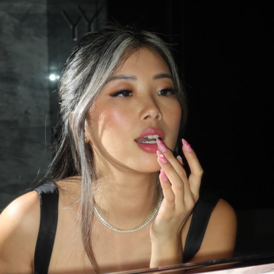 A woman with long dark hair and highlights is applying Glowie By Her's La La La lipstick while gazing into a mirror. She is dressed in a black top and adorned with a pearl necklace. Her salon-quality nails, painted in pink, add an elegant touch as the dimly lit background accentuates her poised presence.