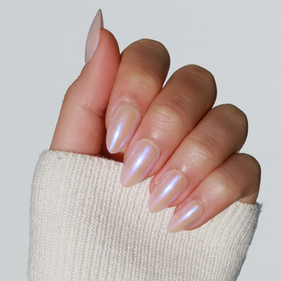 A hand with long, almond-shaped nails adorned in Glowie By Her's "777," showcasing a pearly, iridescent baby pink chrome. The sleeve of a cream-colored knitted sweater partially covers the hand, set against a plain, light-colored background.