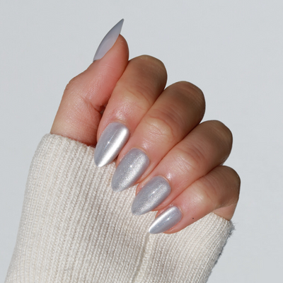 A hand with long, almond-shaped nails painted in the shiny, metallic silver color from the Glowie By Her Velvet Dreams collection (now 20% off). The person is wearing a light beige knitted sweater that partially covers the hand. The background is light grey.
