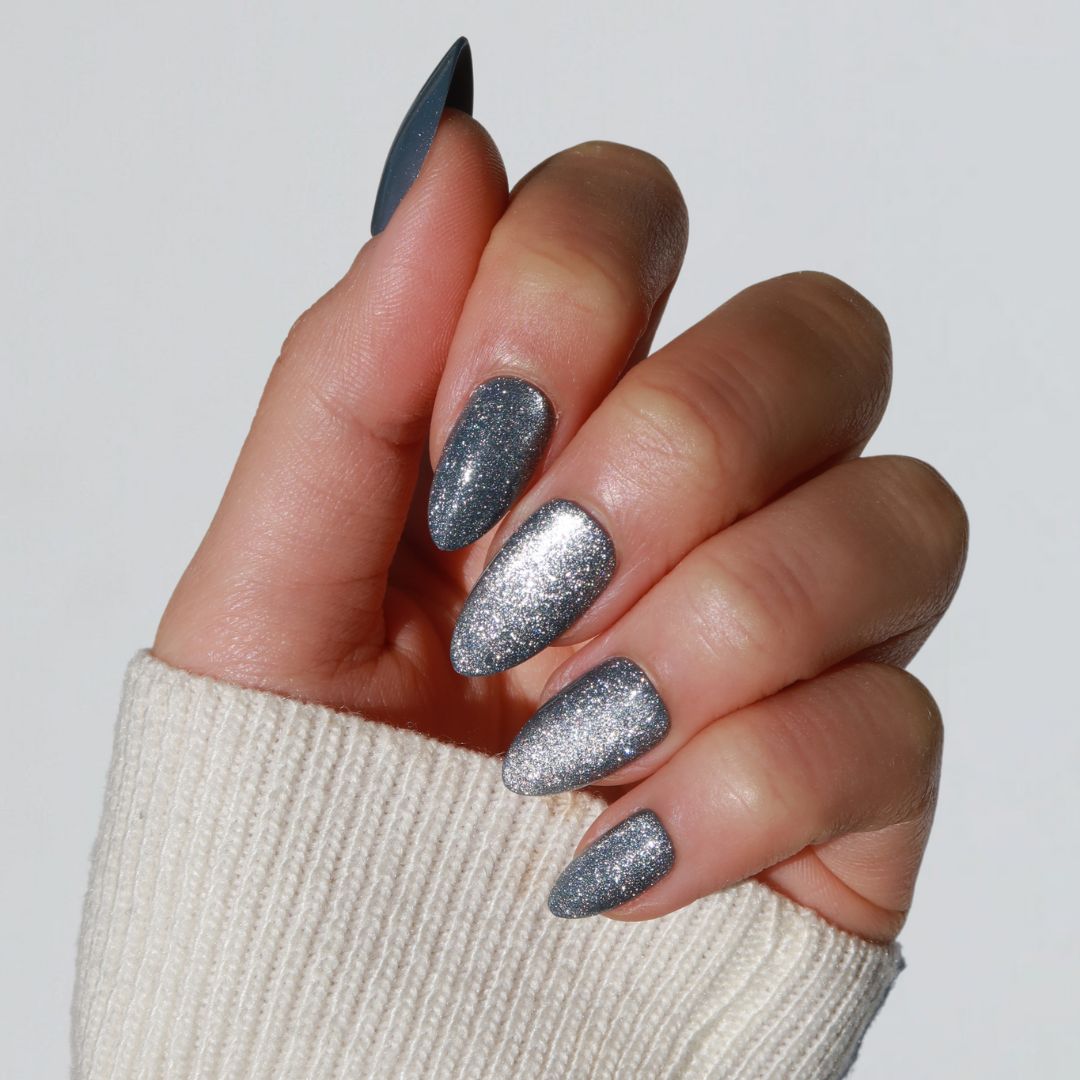 The image showcases a hand with long, almond-shaped nails adorned with Divine, a glittery cat eye silver nail polish by Glowie By Her. The person is wearing a white knitted sweater against a simple light background.