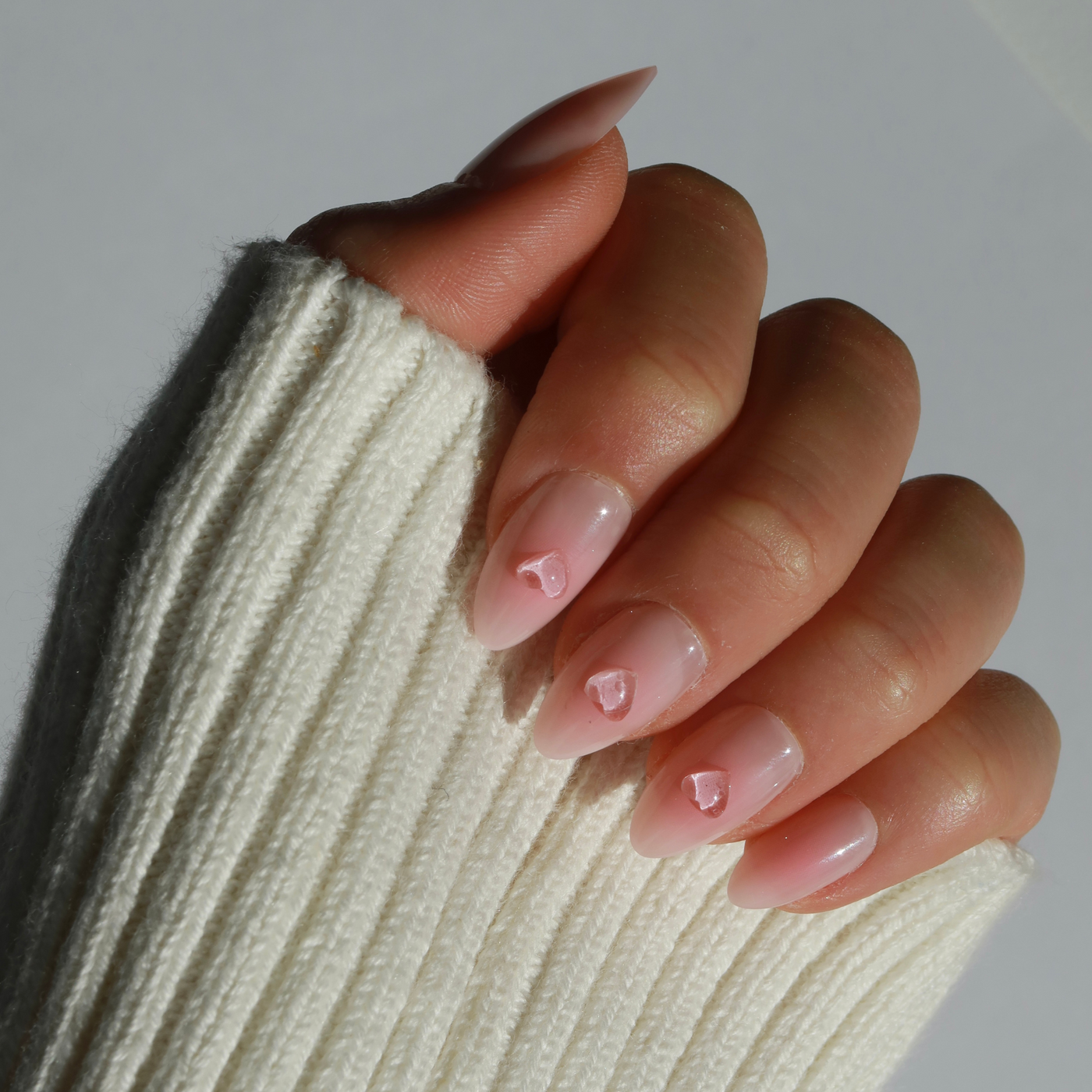 A hand displaying long, almond-shaped nails featuring the "Made You Blush" nail base from Glowie By Her showcases delicate pink rose patterns and delightful 3D gel heart accents. The person, dressed in a cozy white knit sweater, presents their customizable nails against a simple light backdrop.