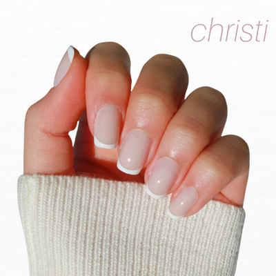 A hand with French tip nails from Glowie By Her's Bridal Bliss (20% Off) collection peeks out from a cream sweater sleeve. The press-on nails are neatly shaped with white tips, set against a plain white background, featuring "christi" elegantly penned in the top right corner.