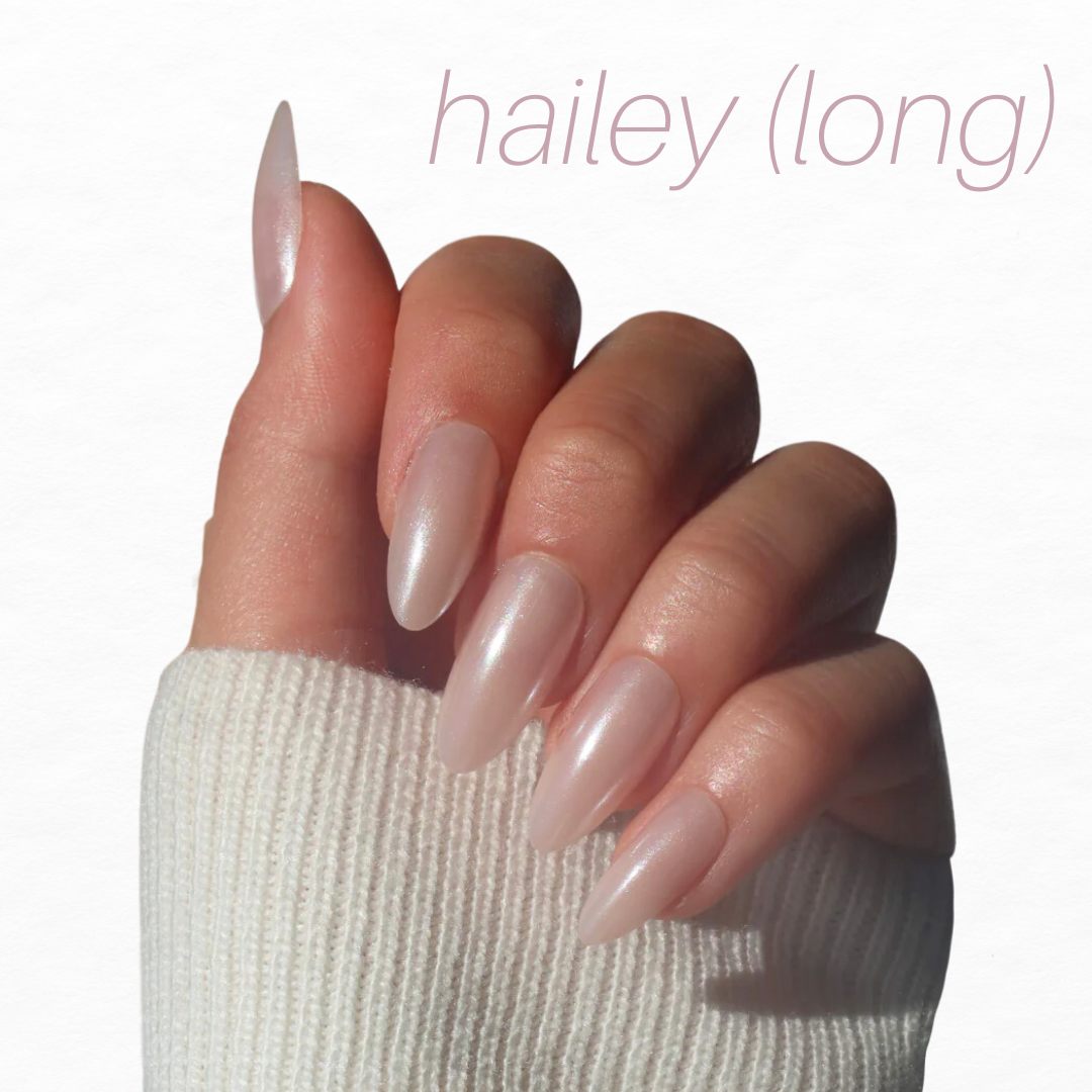 A hand with long, almond-shaped nails showcases pale pink glossy polish and delicate French tips. Partially covered by a white knit sleeve, the text "hailey (long)" in light pink at the top right suggests the elegance of Glowie By Her's Bridal Bliss Collection (20% Off).