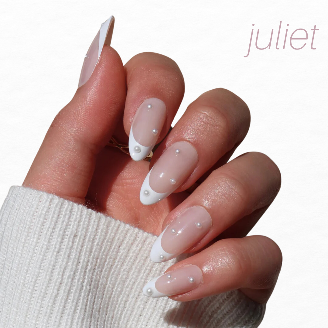 A hand with almond-shaped, press-on nails from Glowie By Her's Bridal Bliss collection features a French manicure with white tips and small pearls. The person dons a white sweater, while "juliet" is elegantly displayed in the top right corner. Enjoy 20% off this exquisite set!.