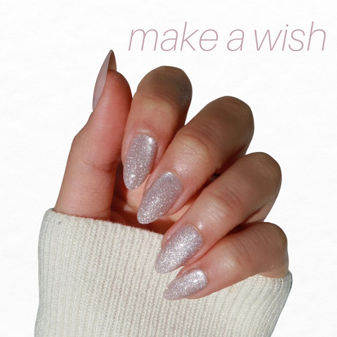 A hand with glittery, silver nails from Glowie By Her's Bridal Bliss (20% Off) collection is positioned against a plain background. The person wears a white sweater. "Make a wish" is written in light pink at the top right corner.