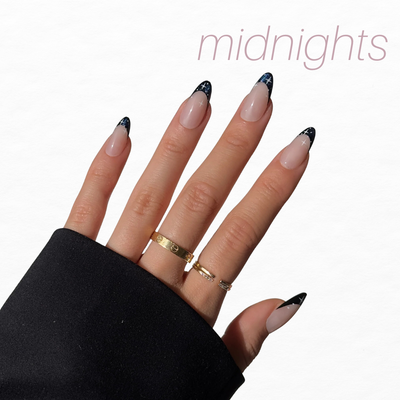 A hand with a black sleeve flaunts almond-shaped nails with black tips and star accents, ideal from Glowie By Her's Glam Night Out (20% Off). "Midnights" appears in light pink above, and the hand is decorated with three gold rings.