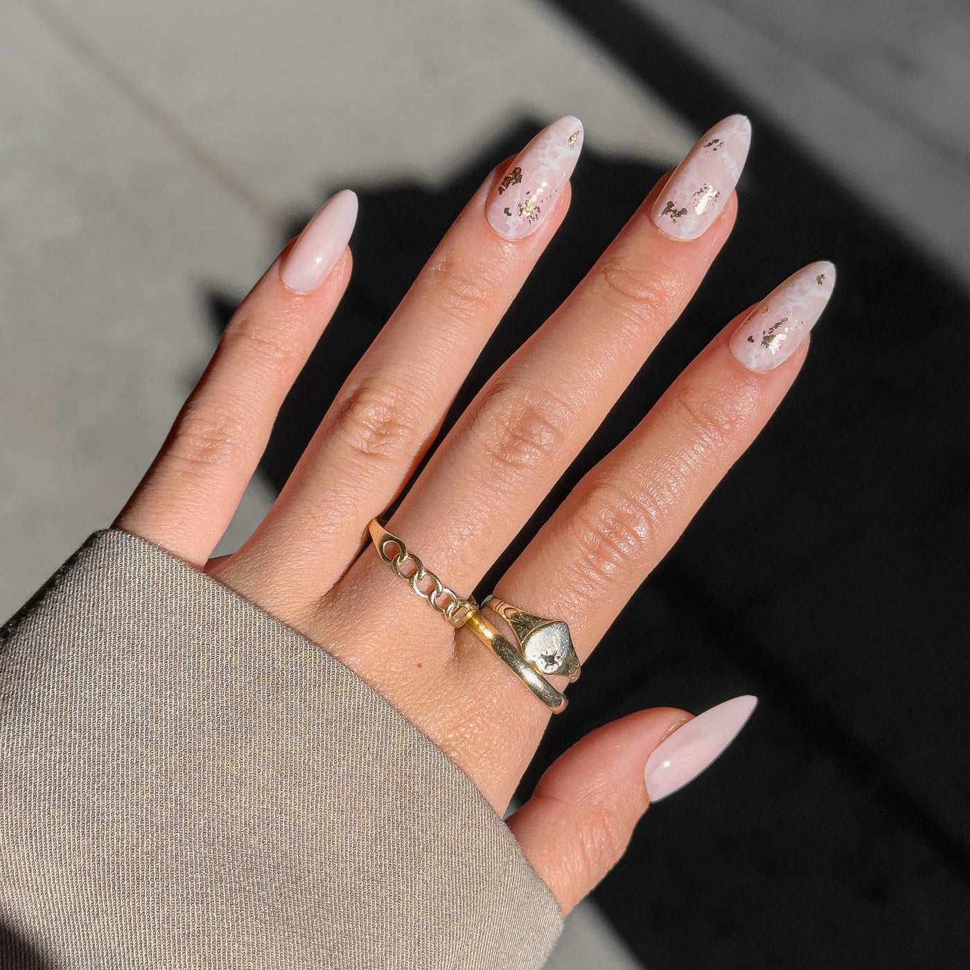 A hand with long, pointed nails displays salon-quality nails in a delicate pale pink and translucent polish embellished with gold flecks. The person is wearing a beige sleeve along with two rings: a gold chain ring and a geometric ring featuring a stone that exudes the elegance of Glowie Co.'s Rose Quartz.