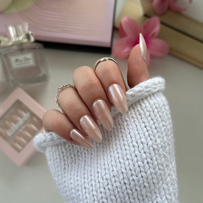A hand with long, salon-quality nails in the shade 'Hailey' from Glowie By Her is wearing a white knitted sleeve. The thumb is lifted slightly, showcasing the shiny finish. Nearby are a perfume bottle, a pink flower, and a box of customizable Hailey nails.