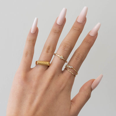 A hand featuring pale pink ombre nails displays three gold rings from Glowie Co.'s Sweetener collection: one engraved with "Amour" on the index finger, another embellished with a small gemstone on the middle finger, and a delicate ring accented with tiny stones on the ring finger.