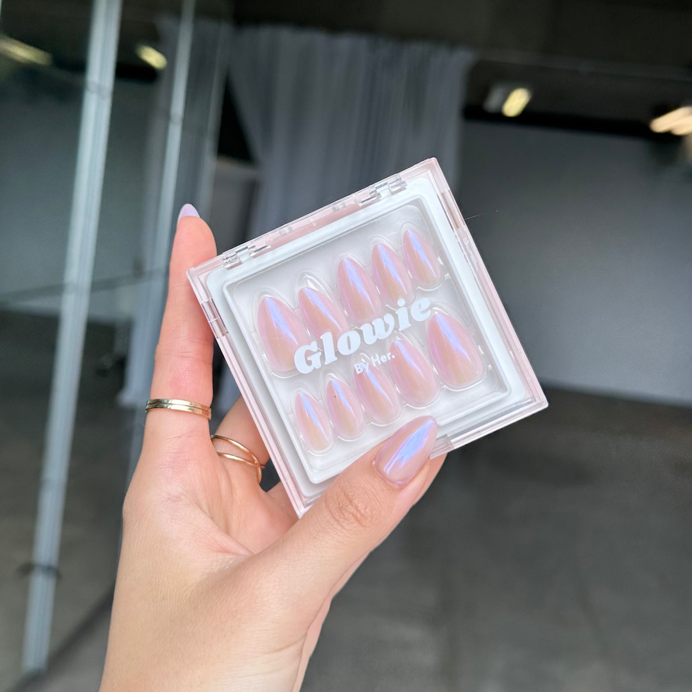 A hand with light purple nails holds a clear box of customizable press-ons from Glowie By Her, labeled "777." The salon-quality nails shimmer with an iridescent baby pink chrome finish against a softly blurred background of white curtains.